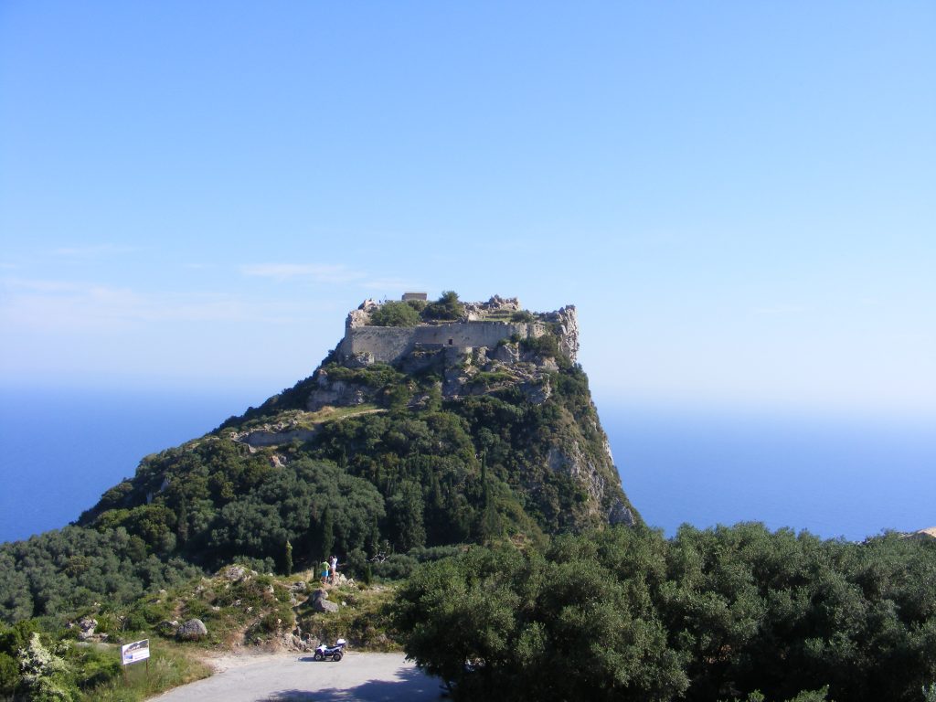 A large castle ruin situated on top of a hill fort overlooking the ocean in Corfu, Greece. The sky is clear blue and the fortress is surrounded by dense tree cover.