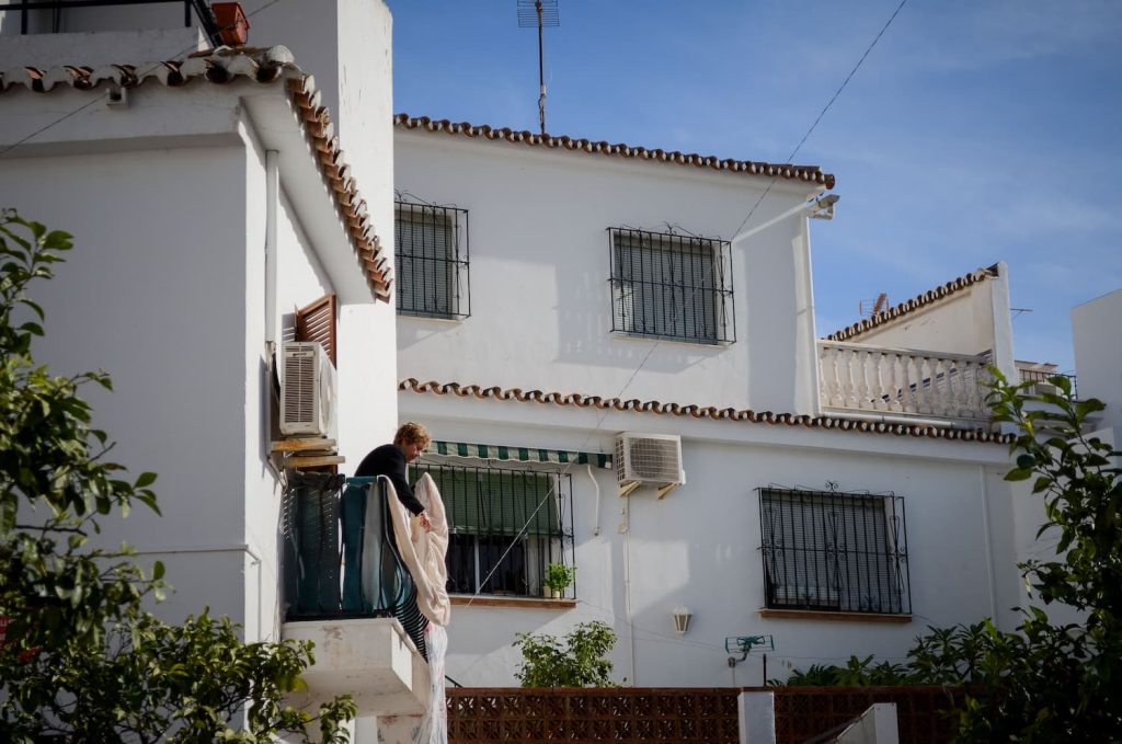A women in Benalmadena Pueblo stands on her balcony hanging her washing out to dry in the sun. The buildings around her are traditional white-washed buildings with terracotta tiled roofs.