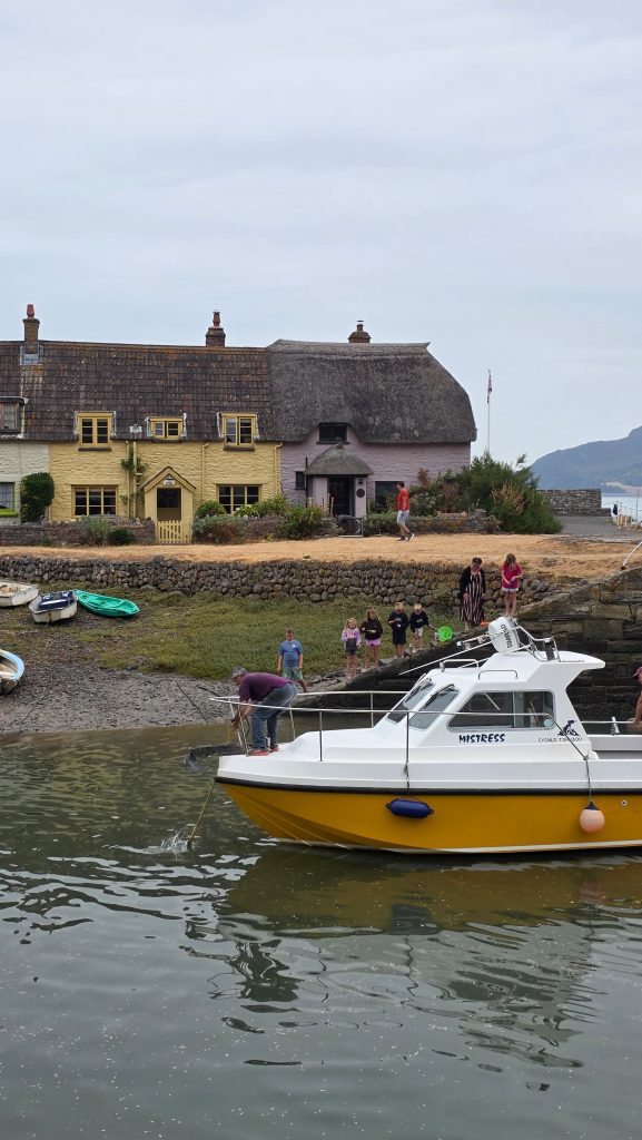 A yellow and white boat sits in Porlock Weir harbour in front of yellow and lilac thatched cottages on a cloudy day.
