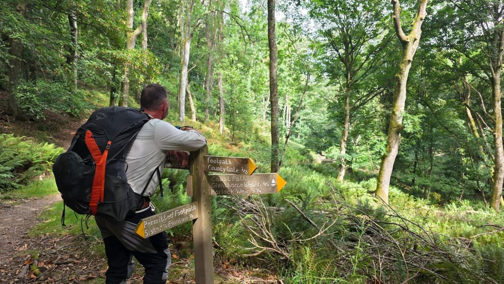Zara's husband Jamie stands leaning on a wooden post in a sunny woodland on the Porlock to Lynmouth hiking trail.