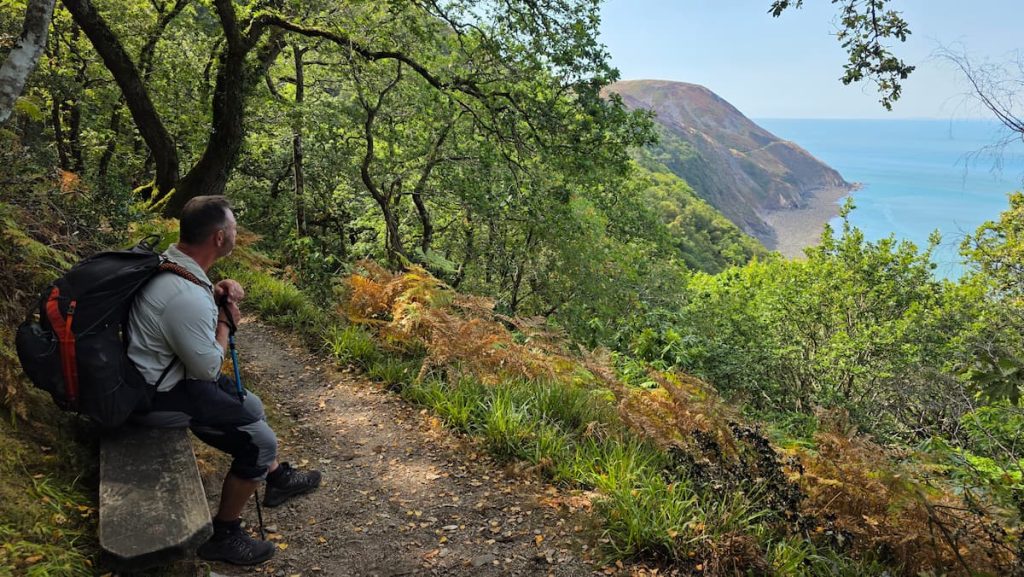 Zara's husband James sits on a wooden bench looking at an incredible coastal view through a gap in the dense treeline on the Porlock Weir to Lynmouth coastal walk.
