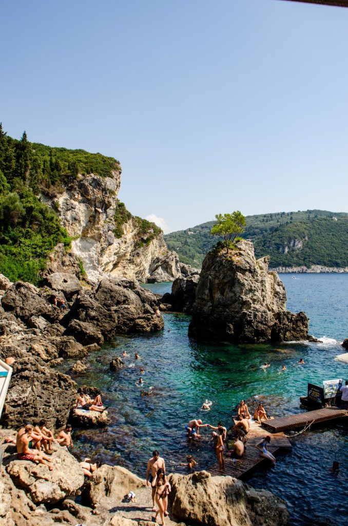 People swim in the blue water at La Grotta Beach Bar in Paleokastritsa Corfu. It's busy with tourists, enjoying the sunshine and views.
