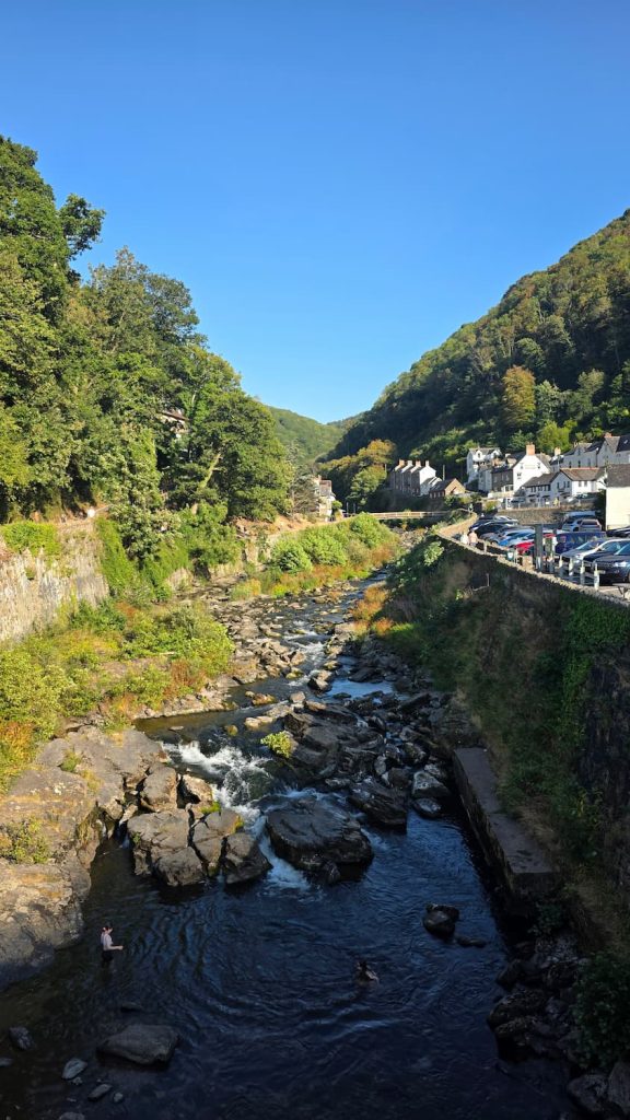 A view looking up the river in Lynmouth. On one side you can see the dense treeline and on the other quaint historical cottages on a sunny day.