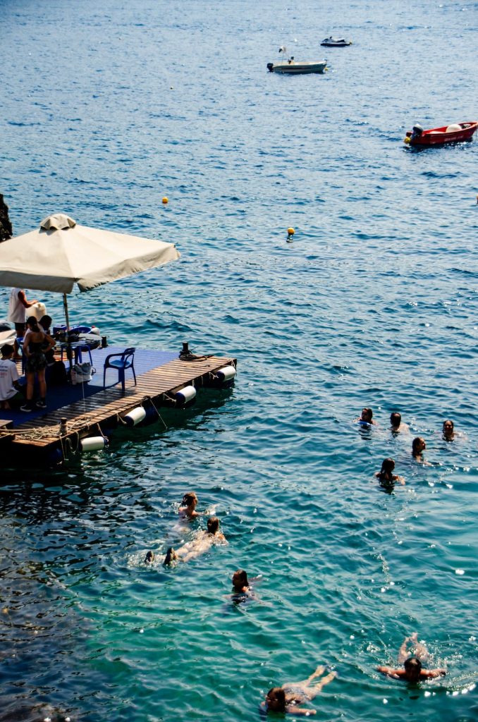 People swim in the blue water at La Grotta Beach Bar in Paleokastritsa Corfu, while others sit on a wooden pontoon under a shaded umbrella.