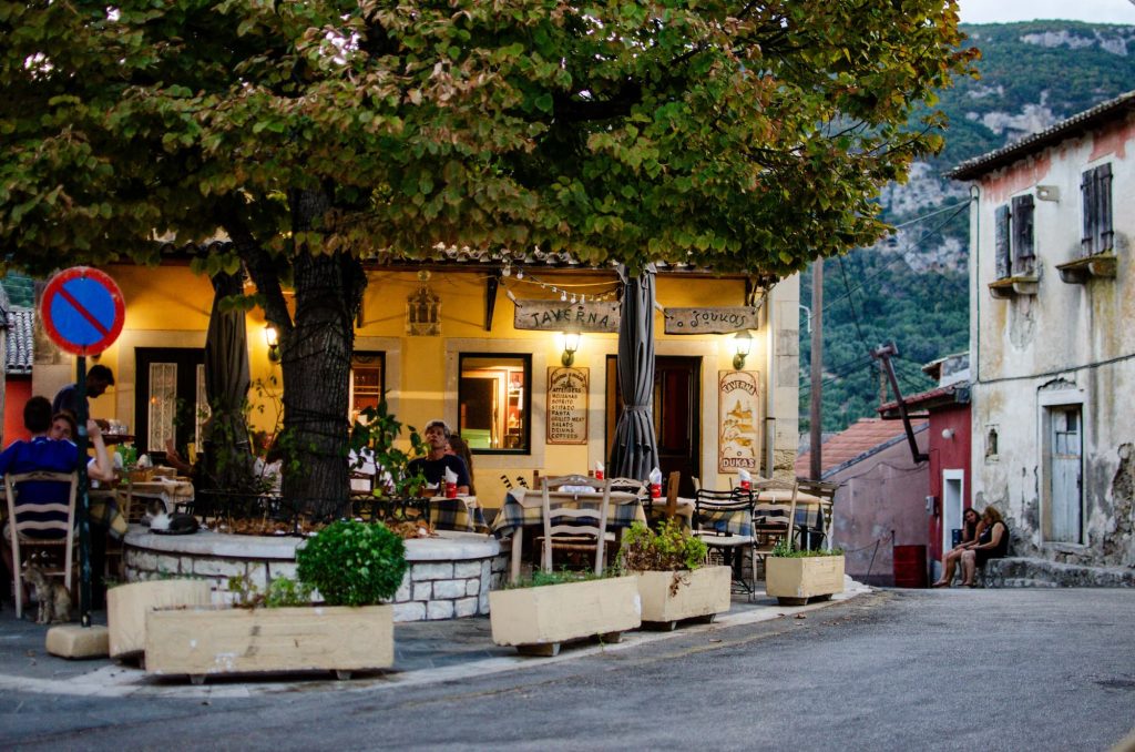The quaint village of Doukades in Corfu at dusk. People sit at a local taverna eating dinner while the taverna is lit up, providing a cosy feel.