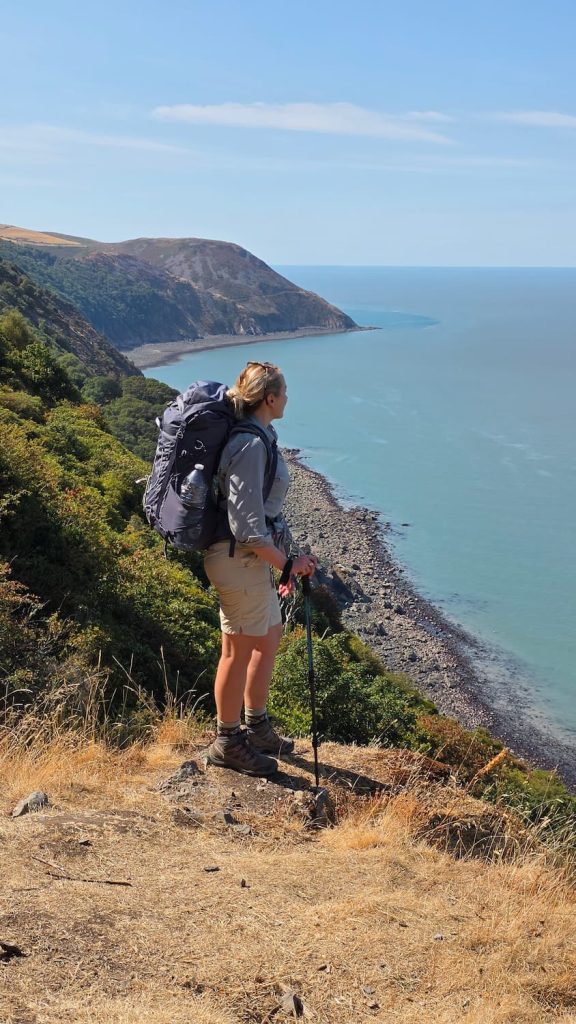 Zara stands at an amazing viewpoint on the South West Coast Path between Porlock Weir and Countisbury. The ocean stretches out in front on a bright sunny day. Zara is wearing hiking clothes and a large backpack.