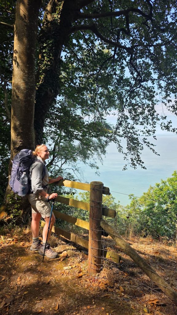 Zara stands at an amazing viewpoint on the South West Coast Path between Porlock Weir and Lynmouth. The ocean stretches out in front on a bright sunny day. Zara is wearing hiking clothes and a large backpack while standing at the viewpoint beneath shaded trees.
