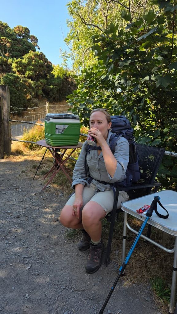 Zara sits on a chair in a shaded spot at an honesty cafe in Gurney's Wood on the Polock Weir to Lynmouth hiking trail. She is sipping a can of Dr Pepper from the honesty cafe.