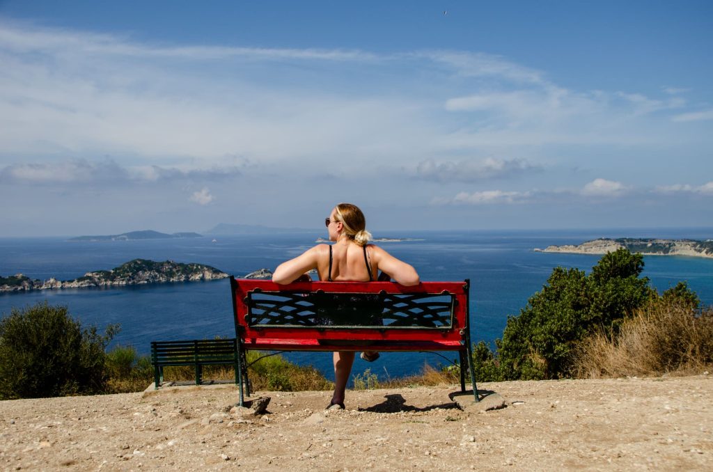 Zara sits on a red bench overlooking incredible ocean scenery in Afionas, Corfu. In the distance, you can see small rugged islands. The sky is partly cloudy, providing an example of what the weather can be like in October.