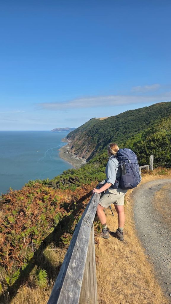 Zara stands at a wooden fence overlooking an amazing coastal viewpoint on a clear sunny day. She si wearing a large backpack and hiking clothes.